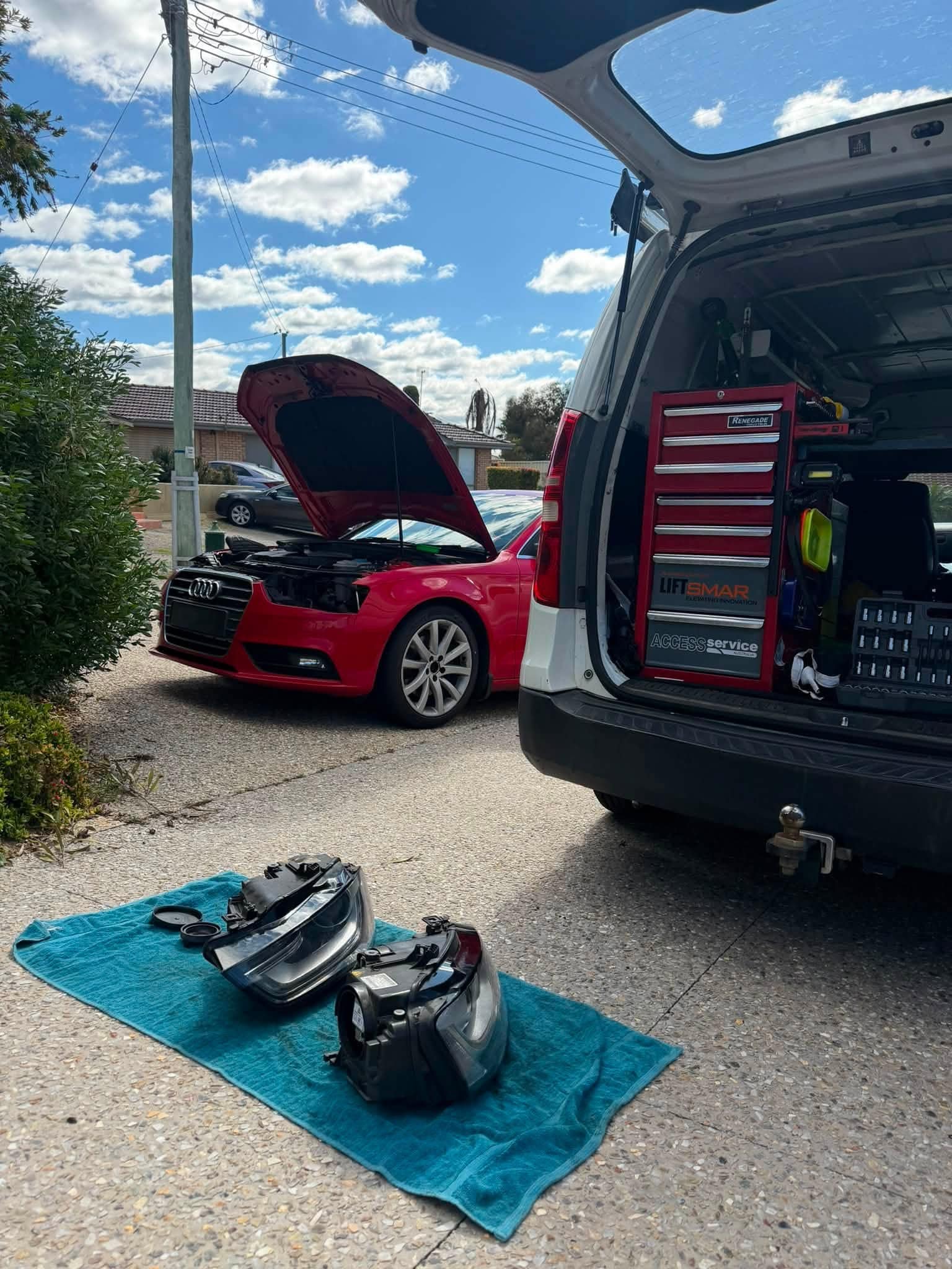 A red Audi parked on a home driveway with its bonnet up and both headlight units laid on a teal towel, with the work van's tailgate open beside it showing a red tool chest.