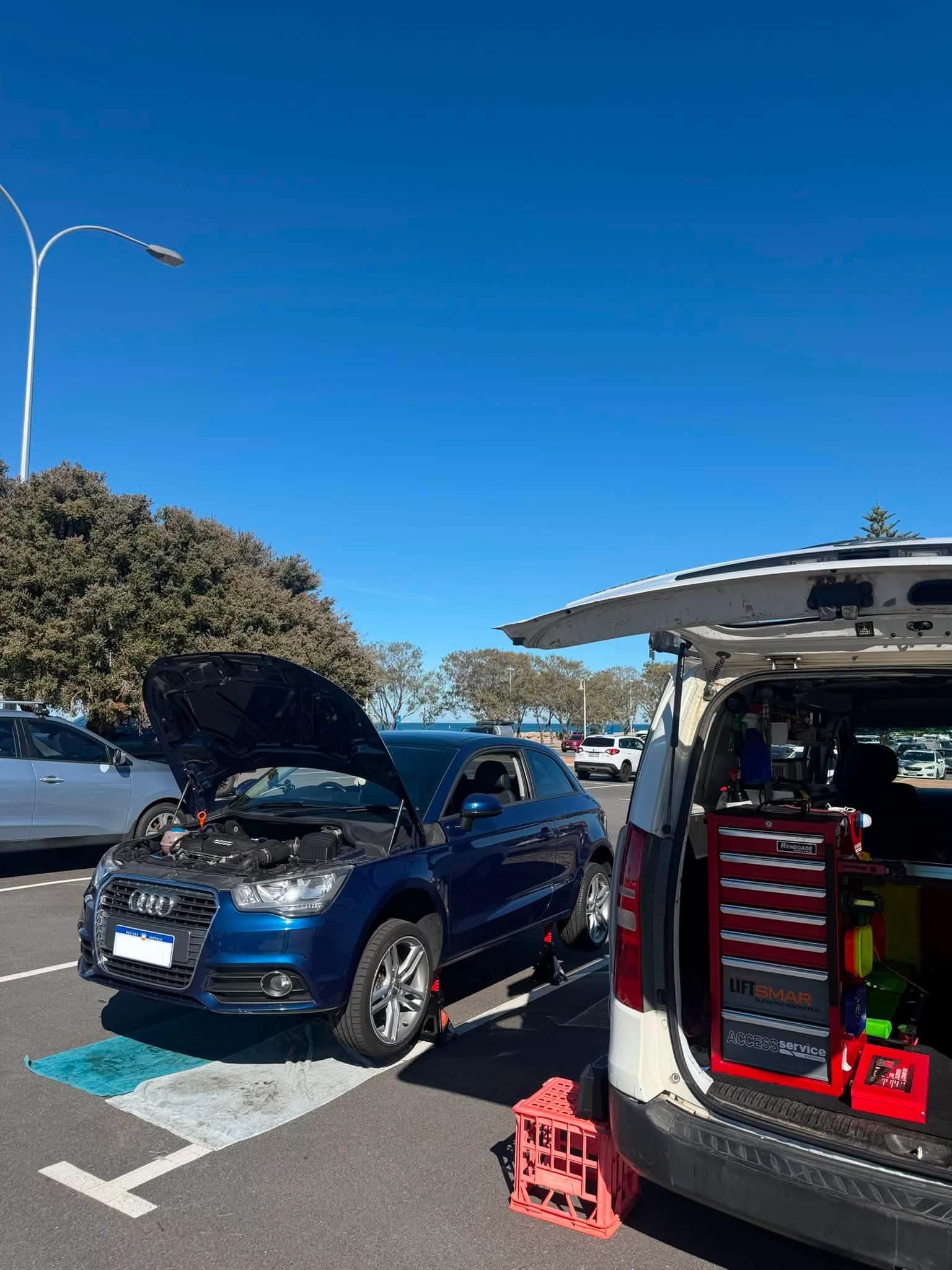 A blue Audi hatchback with its bonnet up in a public car park under a clear blue sky, with the work van and its open tool chest beside it.