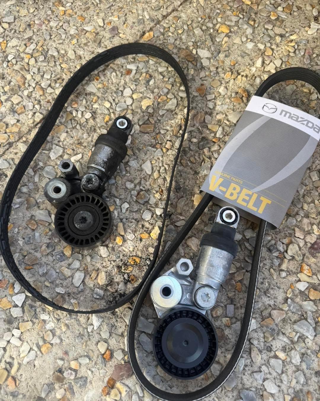 On gravel: an old drive belt and tensioner on the left, and a new Mazda V-belt in its packaging with a tensioner on the right.