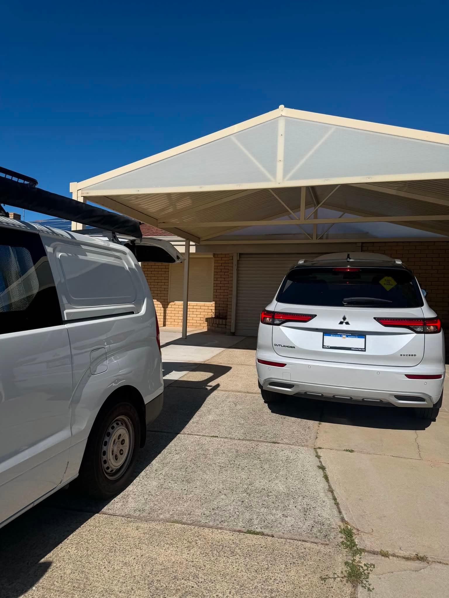 The white work van parked on a sunny driveway beside a white Mitsubishi Outlander under a carport.