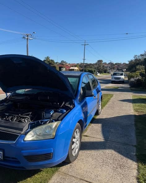 A blue Ford Focus on a suburban street with its bonnet up after breaking down.