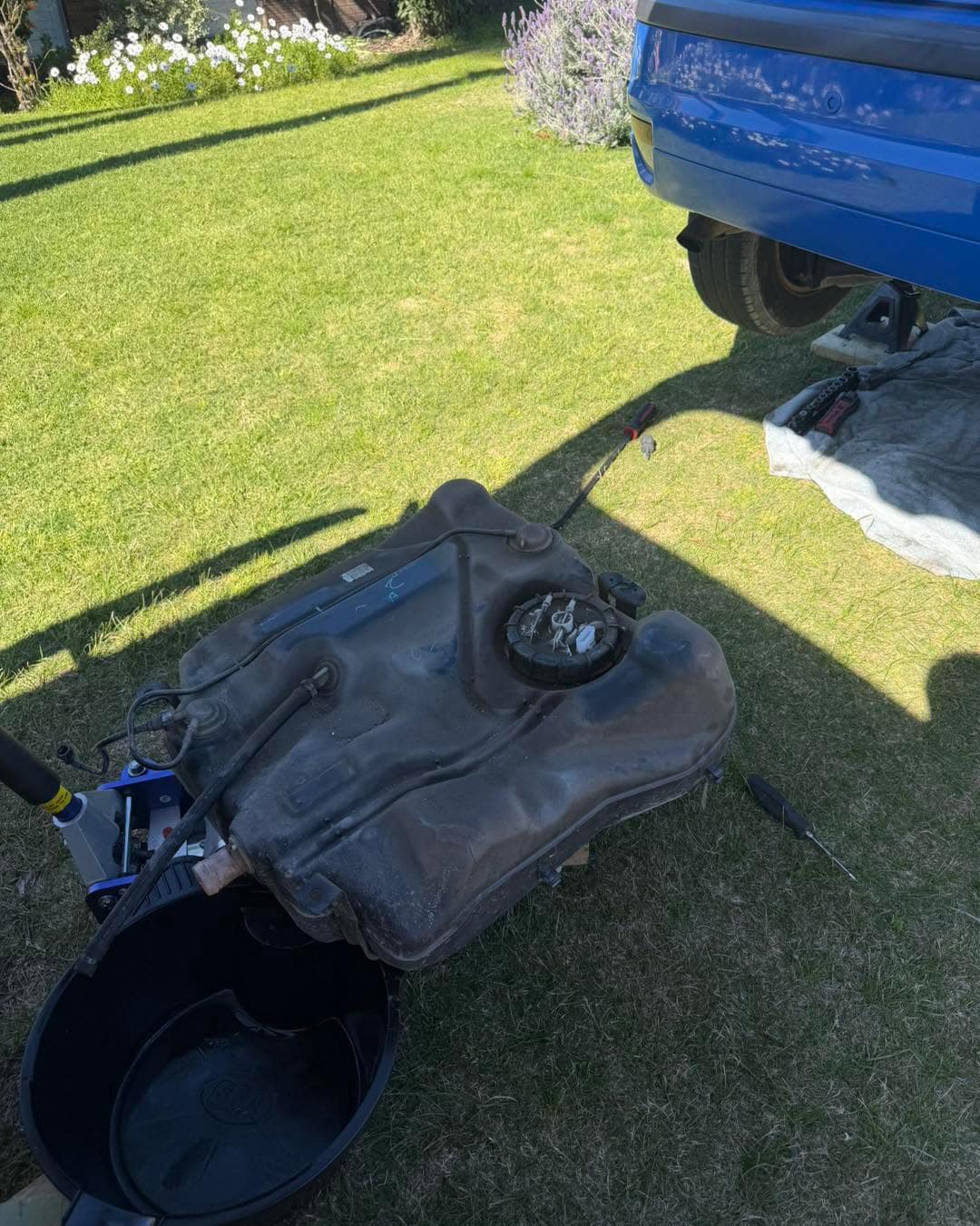 A fuel tank removed from a car and resting on grass beside a drain pan, the blue Focus visible above.
