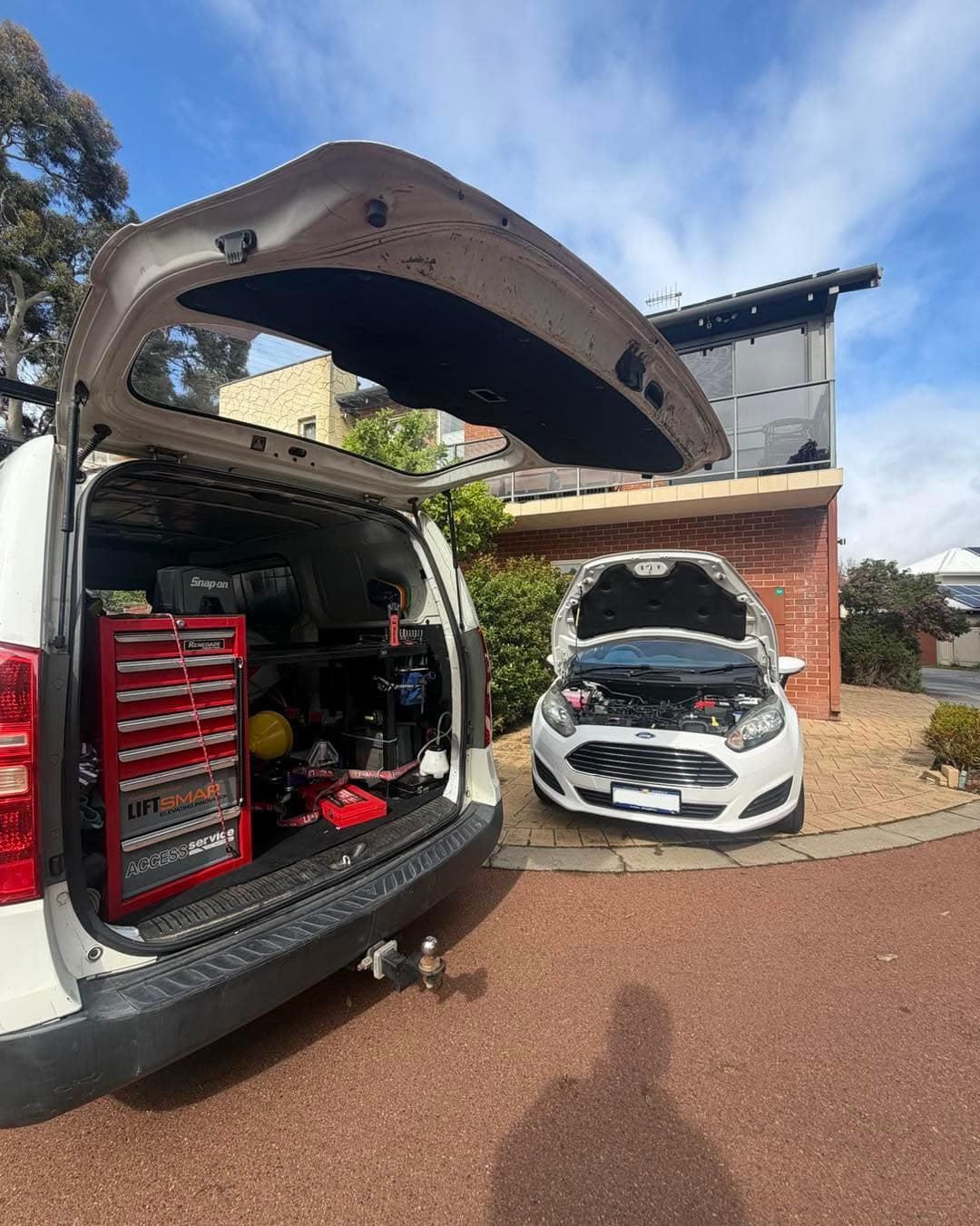 The work van with its tailgate open and tool chest showing, and a white Ford Focus with its bonnet up on a brick-paved driveway.