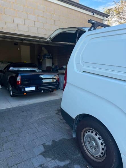 A black Ford ute parked in a home garage, with the white work van pulled up alongside in the foreground.