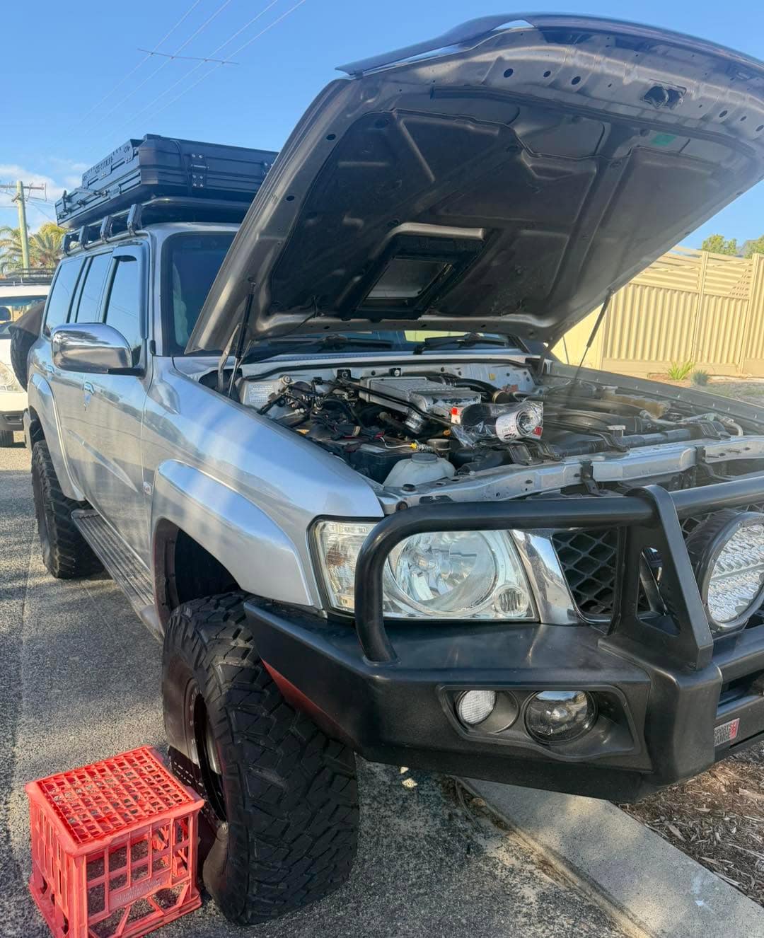 A silver GU Patrol with a bullbar and its bonnet up on a quiet street, a red crate under the front wheel.