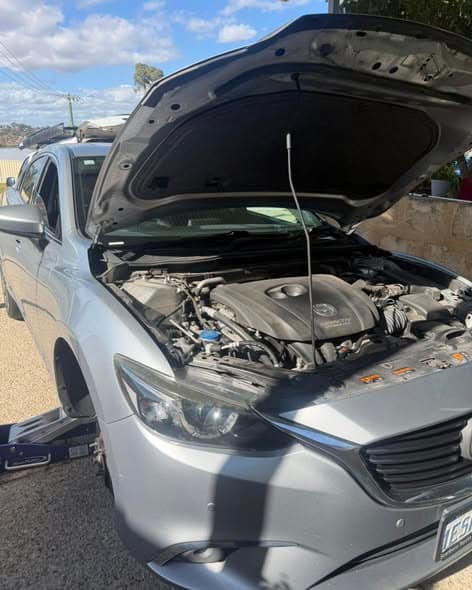 A silver Mazda sedan with its bonnet up beside the kerb, a jack under the front.