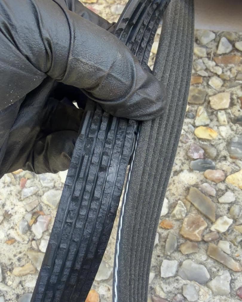 A gloved hand holding a worn ribbed drive belt with crumbling ribs next to a clean one.