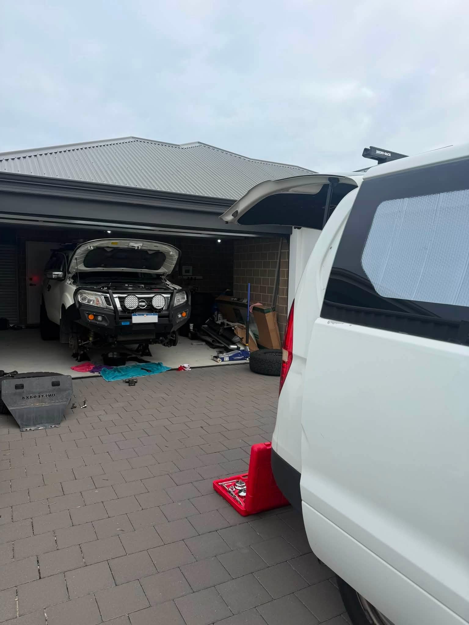 A Nissan Navara with a bullbar and bonnet up inside a home garage, with the work van's open tailgate and a red cone in the foreground.