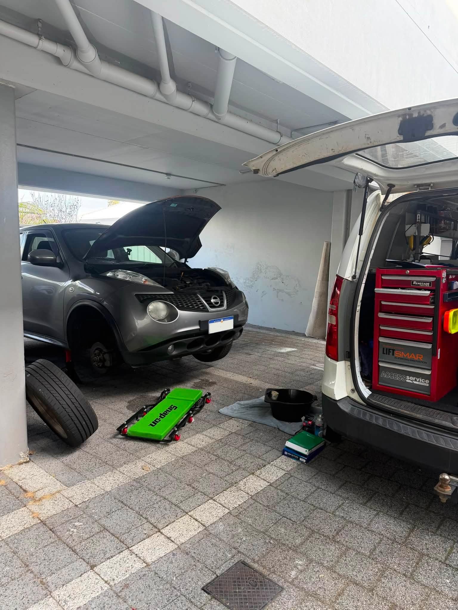 A Nissan Juke in a carport with its bonnet up and a wheel off, a green creeper on the paving and the work van's tool chest open on the right.