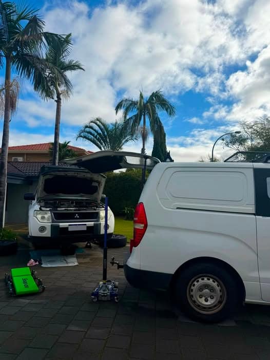 A white Mitsubishi Pajero with its bonnet up and raised on a jack stand on a palm-lined driveway, with the work van and a green creeper alongside.