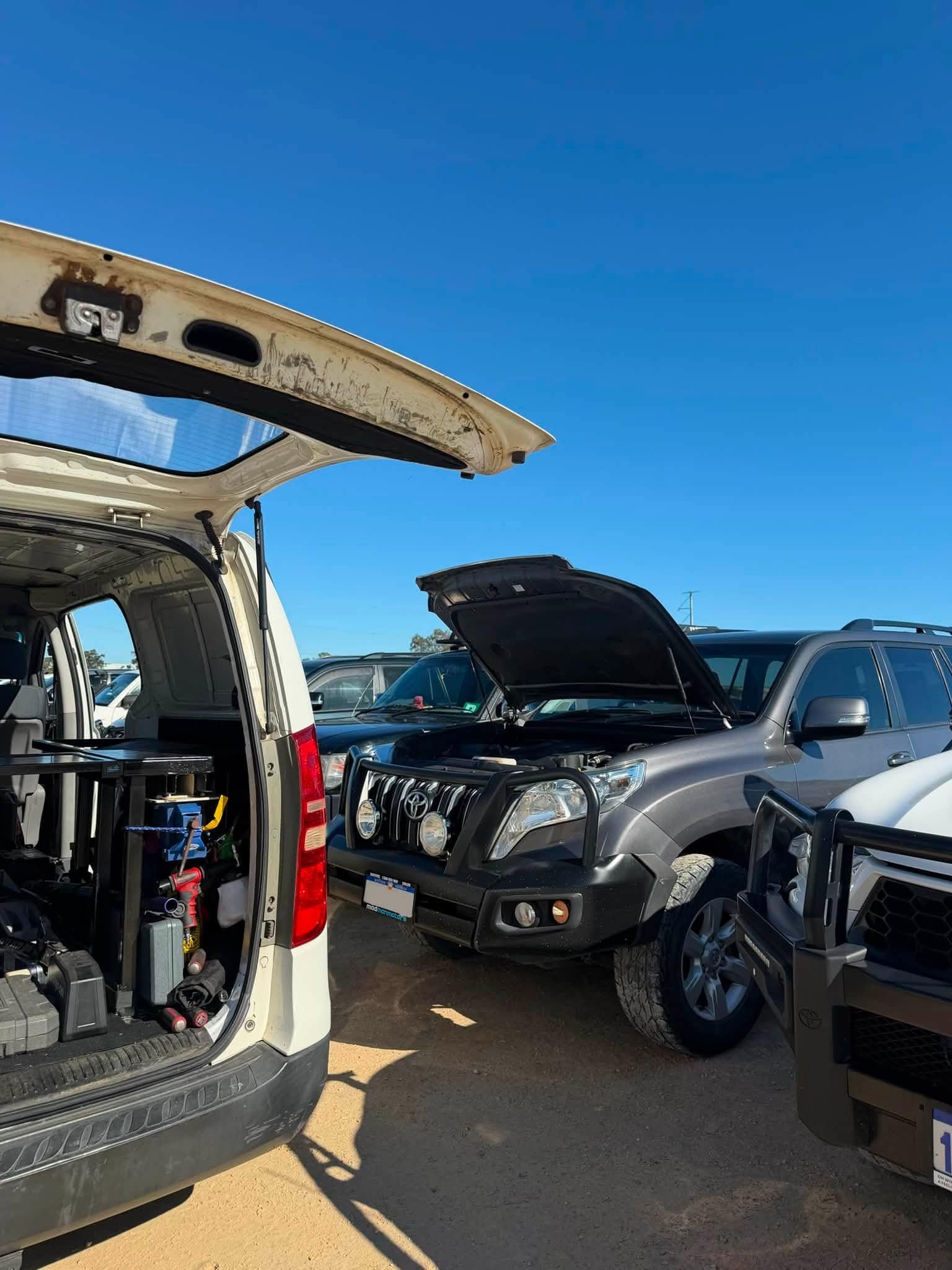 A silver Toyota Prado with its bonnet up in a sunny car park beside the work van with its tailgate open.