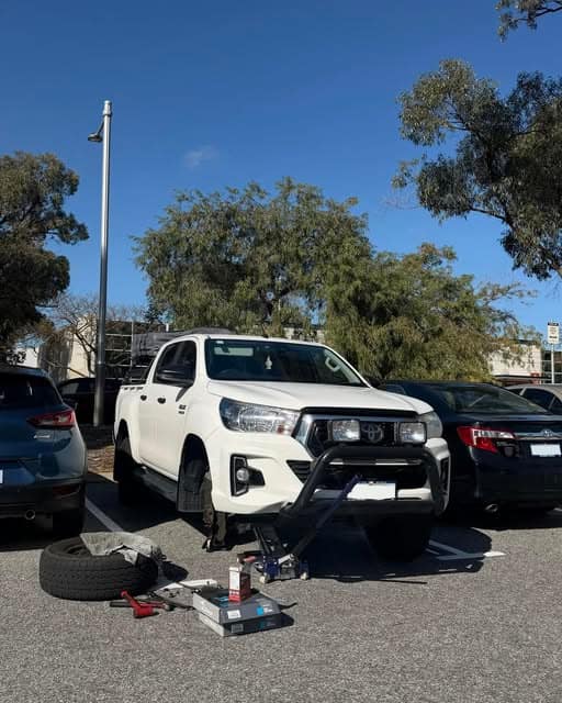A white Toyota Hilux with a bullbar in a car park with a wheel removed and tools, brake parts and oil laid out on the ground in front.