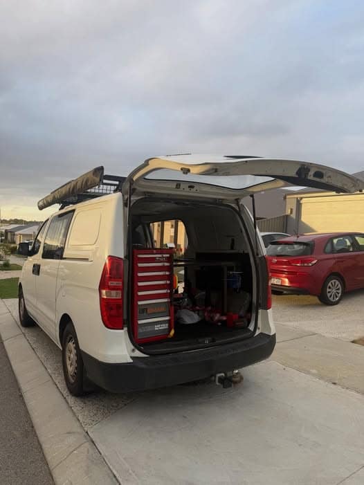 The white work van from behind at dusk with its tailgate open, a roof rack and awning, and a red tool chest in the back.